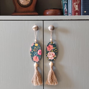 Könnte beinhalten: Zwei ovale Blumenornamente mit Quasten, die an Schrankknöpfen hängen. Die Ornamente sind grün mit rosa und gelben Blumen. Die Quasten sind aus Naturfaser.