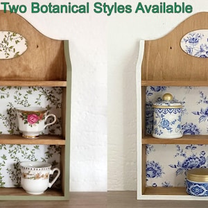 May include: Two wooden wall shelves with arched tops and two tiers. The left shelf features green botanical wallpaper and holds three teacups. The right shelf features blue floral wallpaper and holds a lidded jar and two small containers. Text reads: Two Botanical Styles Available.