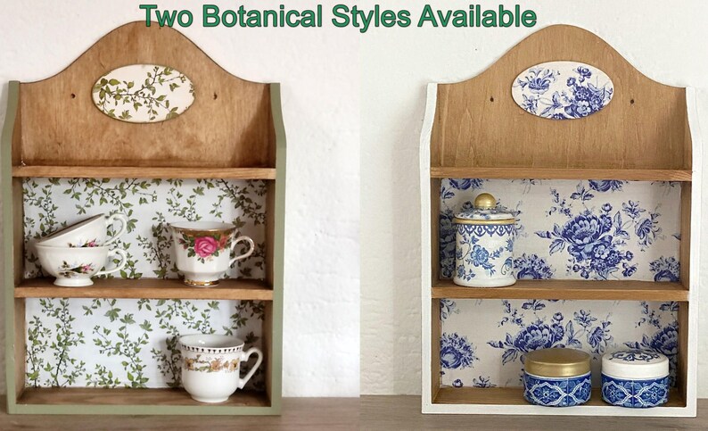 May include: Two wooden wall shelves with arched tops and two tiers. The left shelf features green botanical wallpaper and holds three teacups. The right shelf features blue floral wallpaper and holds a lidded jar and two small containers. Text reads: Two Botanical Styles Available.