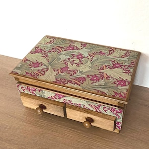 Puede incluir: Una caja de almacenamiento rectangular de madera con dos cajones pequeños y pomos redondos de madera. La tapa superior y el panel frontal sobre los cajones presentan un patrón botánico repetitivo en tonos verde salvia, magenta intenso y crema.