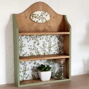 May include: A wooden wall shelf with two tiers and a curved top. The back panel features a white background with a green leaf pattern. An oval plaque with a matching leaf design is centered at the top. The frame is painted a muted sage green.