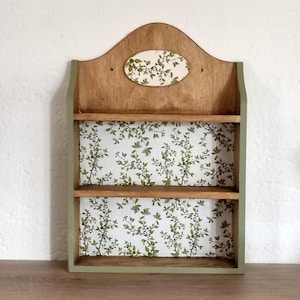 May include: A wooden wall shelf featuring two horizontal shelves and a curved top panel. The back panel and a small oval accent piece at the top are decorated with a white pattern of green leaves and thin brown branches. The frame is painted sage green.