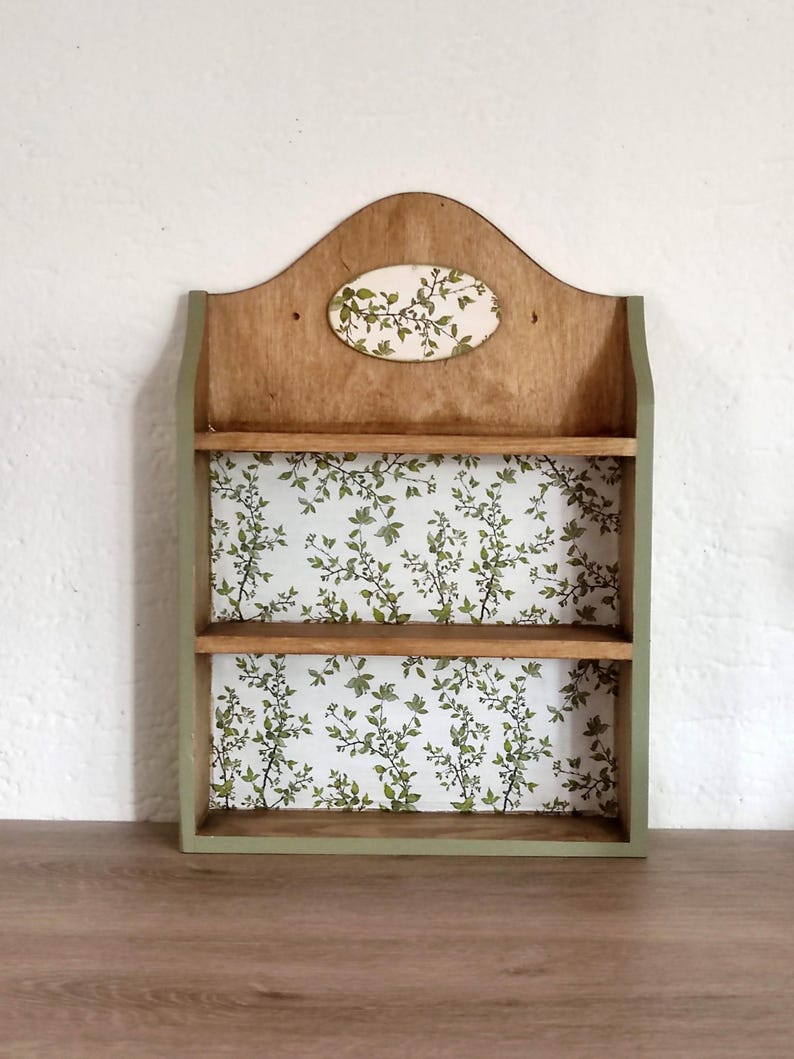 May include: A wooden wall shelf featuring two horizontal shelves and a curved top panel. The back panel and a small oval accent piece at the top are decorated with a white pattern of green leaves and thin brown branches. The frame is painted sage green.