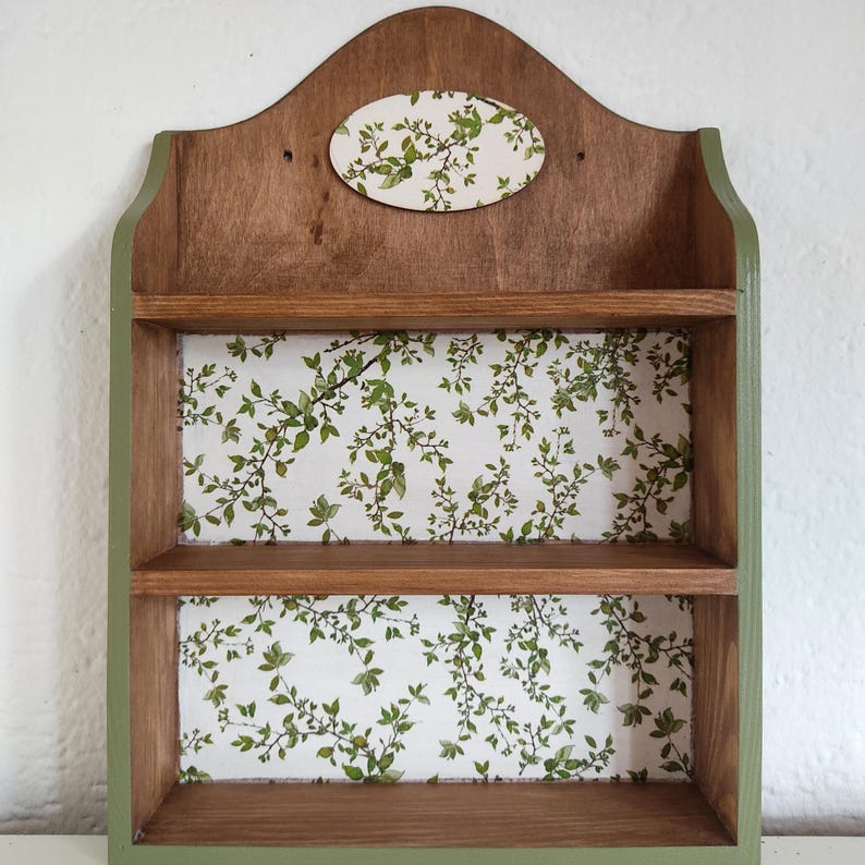 May include: A wall-mounted wooden shelf unit featuring two open tiers and a decorative arched top. The frame is finished in a medium brown wood tone with olive green edges. The back panels are lined with a white paper featuring a green leaf pattern.