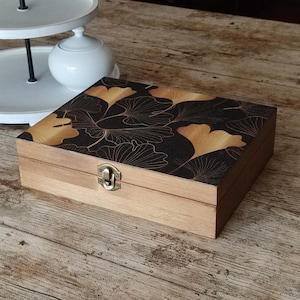 Op de afbeelding: Een rechthoekige houten doos met een natuurlijke lichtbruine afwerking en een scharnierend deksel. De bovenkant van het deksel heeft een zwart oppervlak versierd met een goudkleurig ginkgo-bladpatroon. Een kleine, messingkleurige sluiting.