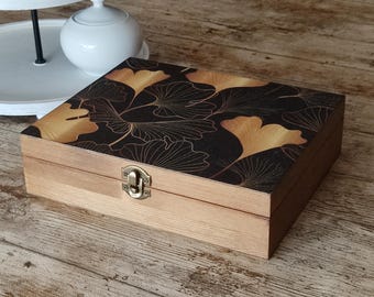 Könnte beinhalten: Eine rechteckige Holzschatulle mit naturfarbenem, hellbraunem Finish und Klappdeckel. Die Oberseite des Deckels ist schwarz und mit einem goldenen Ginkgo-Blattmuster verziert. Ein kleiner, messingfarbener Metallverschluss sichert die Vorderseite.