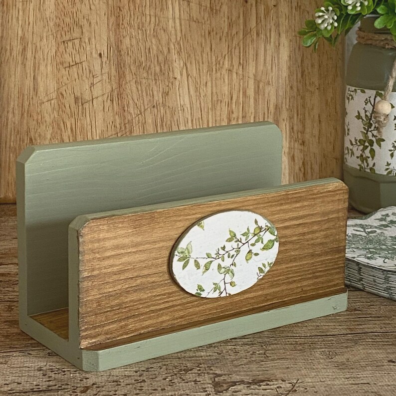 May include: A wooden napkin holder featuring a sage green painted back panel and a natural wood front panel. An oval-shaped insert is centered on the front, displaying a white background with a green botanical leaf branch pattern printed on it.