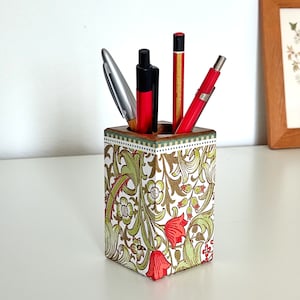 Puede incluir: Un porta lápices de madera con un patrón floral en tonos verdes, marrones y rojos. El porta lápices tiene una forma cuadrada y está lleno de bolígrafos y lápices.
