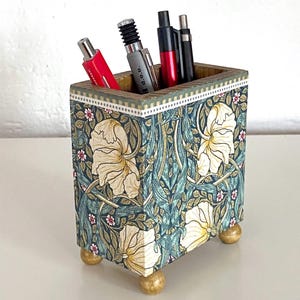 Puede incluir: Un portalápices de madera con un diseño floral en verde azulado, crema y rojo. El portalápices tiene forma cuadrada, se apoya en cuatro pequeños pies dorados y contiene varios bolígrafos y un bolígrafo.