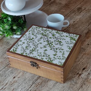 May include: A square wooden box with a hinged lid featuring a white surface decorated with a green leaf and branch pattern. The box has a dark brown wooden frame and a small metal latch on the front for closure, showcasing a natural wood grain finish.