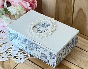 Puede incluir: Una caja rectangular de madera con tapa blanca y un patrón floral azul y blanco en los laterales. La tapa presenta un marco ovalado central con un borde decorativo que encierra un diseño floral a juego con la ilustración de un ave.