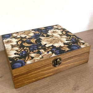 Puede incluir: Una caja rectangular de madera con tapa abatible que presenta un fondo negro decorado con un patrón floral de flores y hojas en tonos crema, beige y azul oscuro. La caja tiene un acabado de veta de madera natural y un cierre de metal cobrizo.