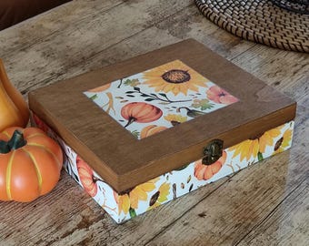 May include: A rectangular wooden box with a hinged lid featuring a metal latch. The lid and sides are decorated with a pattern of orange pumpkins, yellow sunflowers, and green leaves on a white background. The box has a dark brown wood frame.