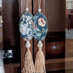 Op de afbeelding: Twee ovale ornamenten met een bloemenprint, houten kralen en beige kwastjes die aan een houten kast hangen.