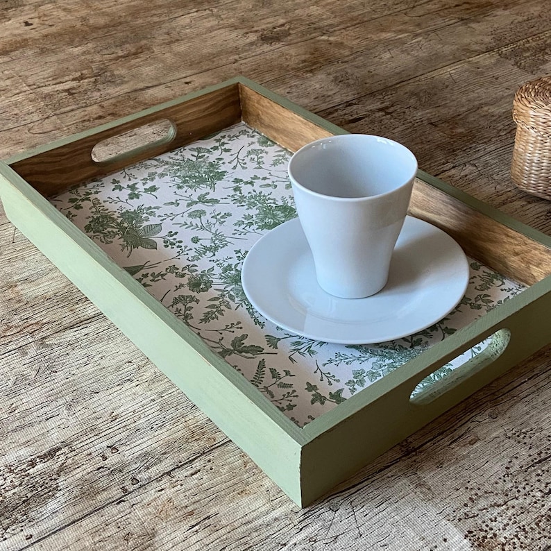 May include: A rectangular wooden serving tray with sage green painted sides and cutout handles. The base features a white surface with a detailed green botanical print. A plain white ceramic cup and matching saucer sit on the tray surface.