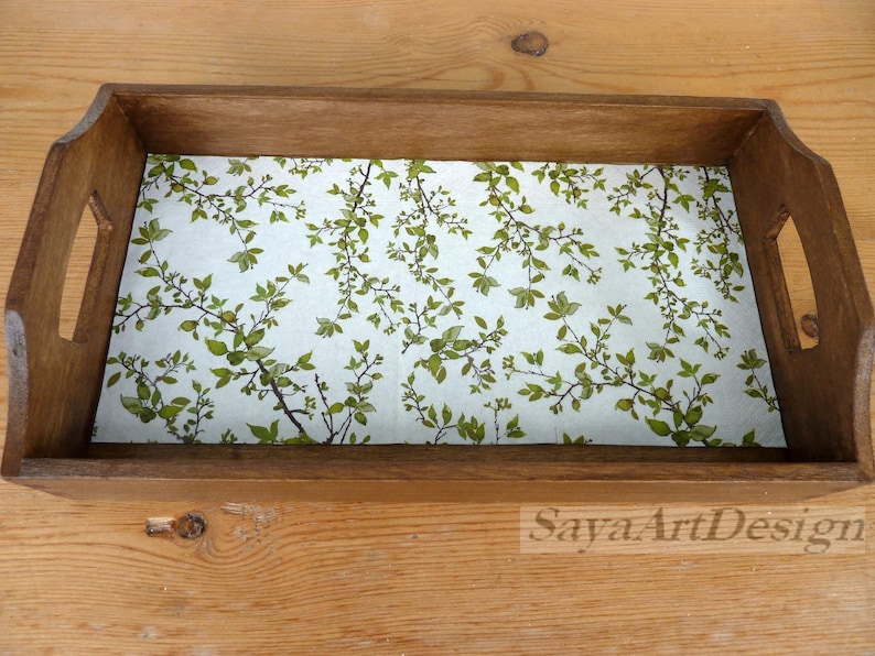 May include: A rectangular wooden serving tray with cutout handles on both sides. The interior base features a light-colored surface decorated with a repeating pattern of green leaves and thin brown branches. The text SayaArtDesign is visible.