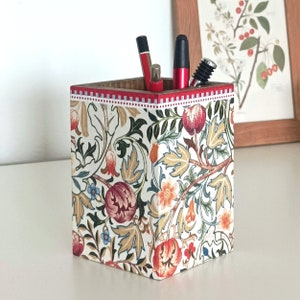 Puede incluir: Un portalápices cuadrado con un estampado floral en rojo, verde y beige. El borde superior está adornado con rojo y blanco. El soporte contiene bolígrafos y un tapón de vino. Una impresión botánica enmarcada está en el fondo.