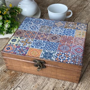 May include: A rectangular wooden box with a hinged lid featuring a multi-colored tile pattern in shades of blue, orange, yellow, and brown. The box has a dark bronze-toned metal latch on the front and a natural wood grain finish on the sides.