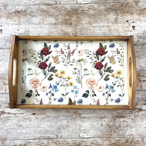 Op de afbeelding: Een rechthoekig houten dienblad met een lichtbruine afwerking en geïntegreerde handgrepen aan de zijkanten. De bodem heeft een witte achtergrond versierd met een herhalend patroon van kleurrijke wilde bloemen, waaronder rode rozen, gele madeliefjes en oranje vlinders.