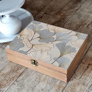 Può includere: Una scatola rettangolare in legno con coperchio incernierato caratterizzata da un motivo stampato di foglie di ginkgo azzurre e dorate su sfondo crema. La scatola presenta una chiusura metallica color oro sul davanti e finitura in legno.