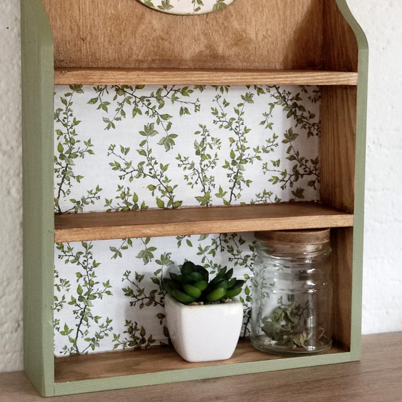May include: A wall-mounted wooden shelf unit featuring two tiers with a natural wood finish and sage green painted sides. The back panels are decorated with a white wallpaper pattern of green leaves and thin brown branches. A small white pot with a green succulent and a clear glass jar with a cork lid sit on the bottom shelf.