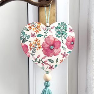 Peut inclure: Une décoration en bois en forme de cœur blanc avec un motif floral dans des tons de rose, orange et vert. La décoration a un pompon bleu clair et une perle en bois.