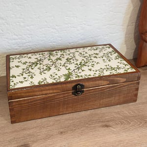 May include: A rectangular wooden box with a dark brown stained finish and a hinged lid. The top of the lid features a cream-colored panel decorated with a green leafy vine pattern. A small, dark metal latch is centered on the front of the box.