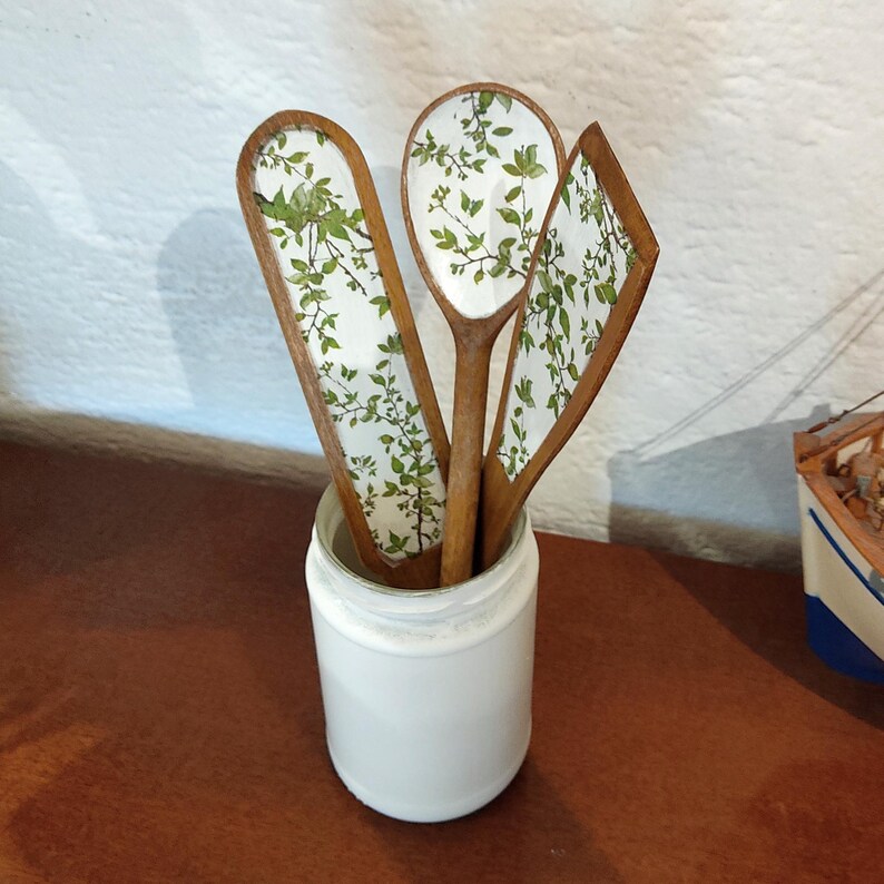 May include: Three wooden kitchen utensils with a white background and a green leaf pattern on the front, including a spoon, a flat spatula, and a slanted spatula, all standing upright inside a plain white cylindrical container.