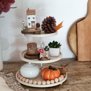 May include: A three-tiered white metal tray stand decorated with seasonal items. The top tier holds a miniature white house, a pinecone, and a leaf. The middle tier features wooden slices, two mushroom figurines, and a succulent. The bottom tier has a white lidded jar, an orange pumpkin, and wooden beads.