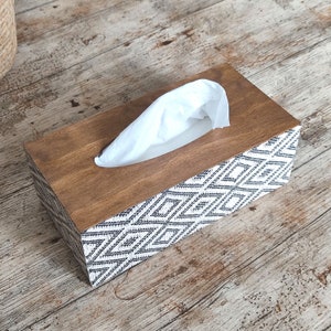May include: A rectangular tissue box cover featuring a smooth, dark brown wooden top and sides decorated with a black and white geometric diamond pattern. A single white tissue is pulled through the oval opening in the center of the wooden lid.