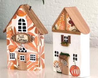 May include: Two miniature wooden house figurines. The left house features an orange and brown leaf pattern with a sign reading Fall Cottage. The right house is white with a brown roof, a painted orange pumpkin, and a small window with shutters.