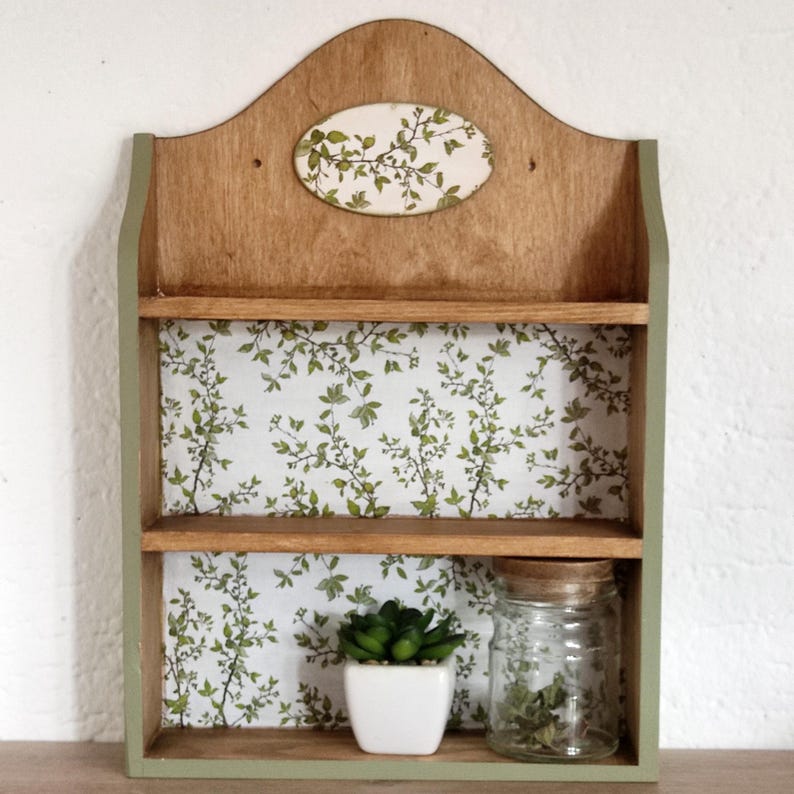 May include: A wooden wall shelf with two tiers, featuring a light brown wood finish and sage green painted sides. The back panel is decorated with a white and green botanical leaf pattern. An oval plaque with the same leaf design is at the top.
