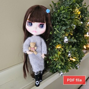 May include: A doll with long brown hair wearing a gray knitted dress and black and white checkered leggings. The doll is holding a small brown teddy bear. The doll is standing on a shelf with a green wreath behind it. The text "PDF file" is visible in the lower right corner of the image.