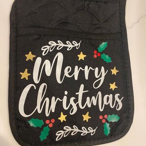 May include: Black pot holder with the words "Merry Christmas" in white script. The design includes gold stars, green holly leaves, and red berries. A loop is attached for hanging.