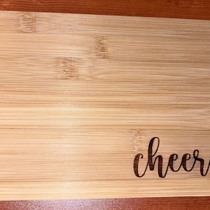 May include: A rectangular bamboo cutting board with rounded corners. The board has a natural wood grain pattern and the word "cheers" is etched in a cursive font on the lower right corner.