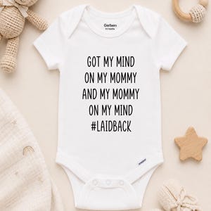 May include: White baby onesie with the text "GOT MY MIND ON MY MOMMY AND MY MOMMY ON MY MIND #LAIDBACK". The onesie is surrounded by baby items, including a teddy bear, wooden star, and knitted booties.