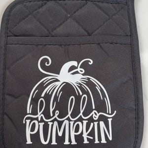 May include: Black quilted oven mitt featuring a white pumpkin design and the words "Hello Pumpkin". The mitt has a hanging loop. A seasonal kitchen accessory.