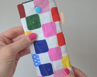 May include: A colorful, rectangular glasses case with a patchwork design of squares in red, pink, blue, green, and yellow. The case has a blue button closure and a small, peach-colored tag with the word "bdoo".