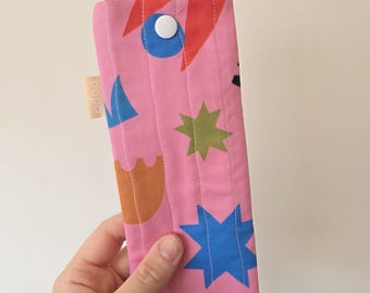 May include: A pink, quilted glasses case with a colorful geometric pattern. The case features a button closure and the word "oopa" on a small tag. The design includes blue stars, orange shapes, and green accents.