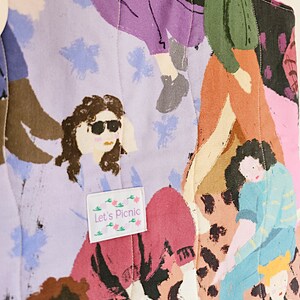 Pu&ograve; includere: Una borsa tote trapuntata con una stampa colorata di figure in varie pose. Il design include sfumature di viola, rosa, marrone e blu, con un'etichetta che recita "Let's Picnic". La borsa presenta un motivo cucito.