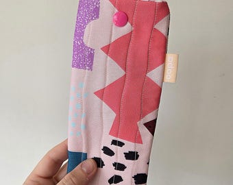 May include: A colorful, quilted glasses case with a pink button closure. The case features a patchwork design with pink, purple, and black geometric shapes. A small, rectangular tag with the text "bcloo" is attached to the side.