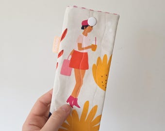 May include: A cream-colored glasses case with a button closure. The case features a colorful illustration of a person holding a drink and shopping bag, with yellow flowers and red leaf accents. The tag reads "oopa".