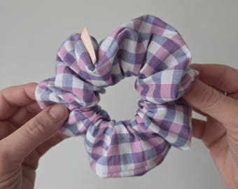 May include: A fabric hair scrunchie featuring a purple, white, and grey gingham check pattern. The scrunchie has a gathered, ruffled texture and includes a small, light-colored rectangular fabric tag with partially visible text attached to the side.