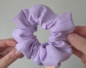 May include: A light purple fabric scrunchie with a gathered, ruffled texture. The hair accessory features a small, rectangular peach-colored fabric tag attached to the outer edge with the lowercase white text oopa printed on it.