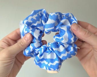 May include: A blue and white patterned scrunchie. The fabric features a repeating wavy design in a bright blue color against a white background. The scrunchie is gathered and appears soft and flexible, suitable for holding hair.
