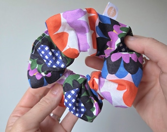 May include: A colorful scrunchie with a variety of floral and geometric patterns in orange, purple, blue, and green. The scrunchie is held in a person's hands. A small tag with the word "cope" is attached to the scrunchie.