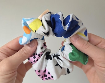 May include: A colorful scrunchie with a white base and abstract patterns in blue, yellow, pink, green, and black. The fabric is gathered around an elastic band, creating a textured, circular shape. A fun hair accessory.