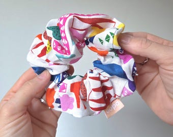 May include: A colorful scrunchie with a white background and a vibrant pattern of red, pink, yellow, blue, and green shapes. The scrunchie is held between two hands. A small tag with the word "oopa" is visible.