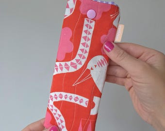 May include: A rectangular, quilted glasses case with a vibrant red background and a repeating pattern of pink and white shapes. The case has a pink button closure and a small tag with text. The interior lining is a patterned pink and purple fabric.