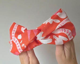 May include: A vibrant orange headband with a white and pink geometric pattern. The fabric is twisted at the center, creating a stylish accessory. The headband is held up by two hands.